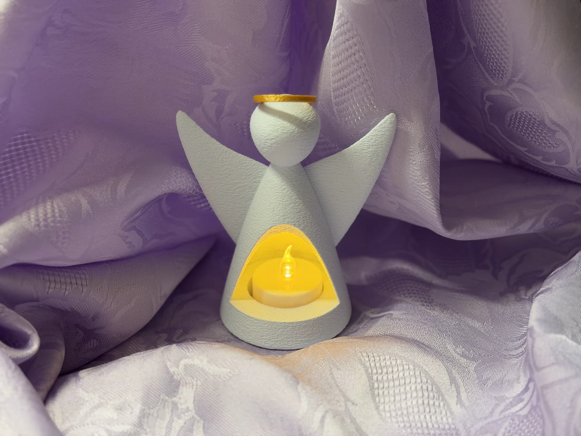 Anjo porta-vela artesanal branco com auréola dourada e vela tealight acesa, fundo tecido lilás suave, vista frontal