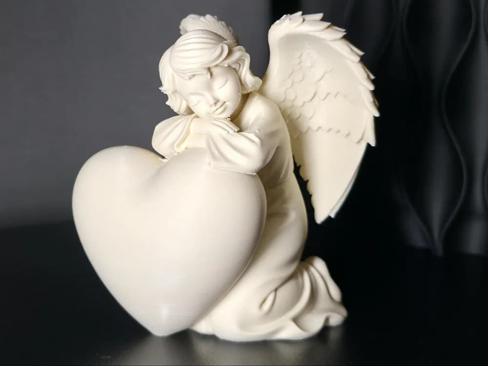 Vista aproximada do anjo artesanal branco pousado sobre coração, iluminação suave realçando detalhes das asas e do rosto