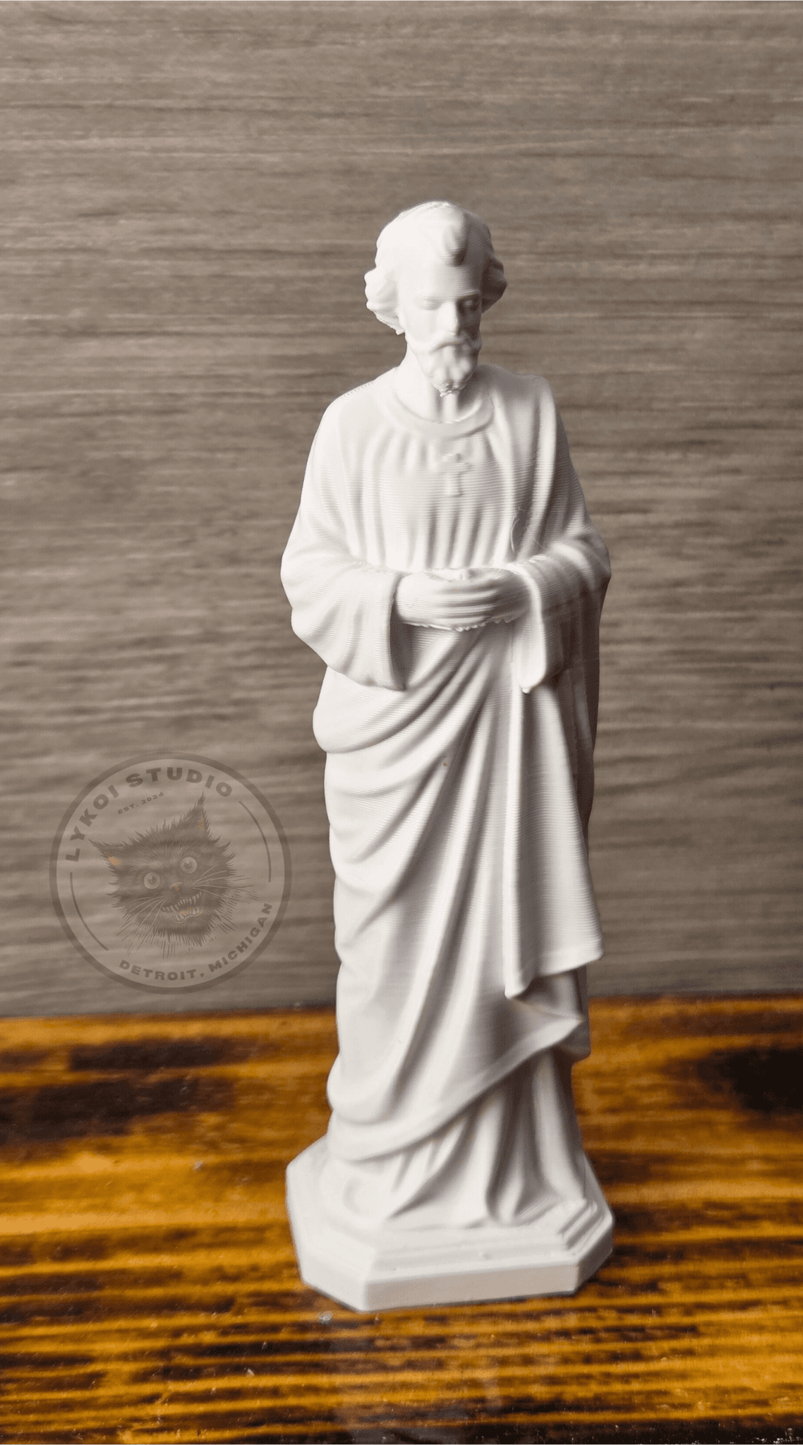 Escultura de São José sobre superfície de madeira com parede cinza ao fundo, detalhes das vestes e postura